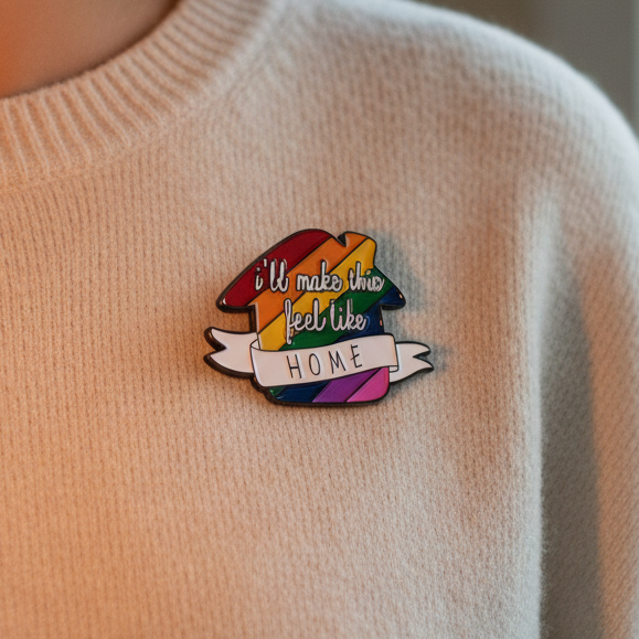 Enamel pin reading “I'll make this feel like HOME” on a rainbow-striped house design