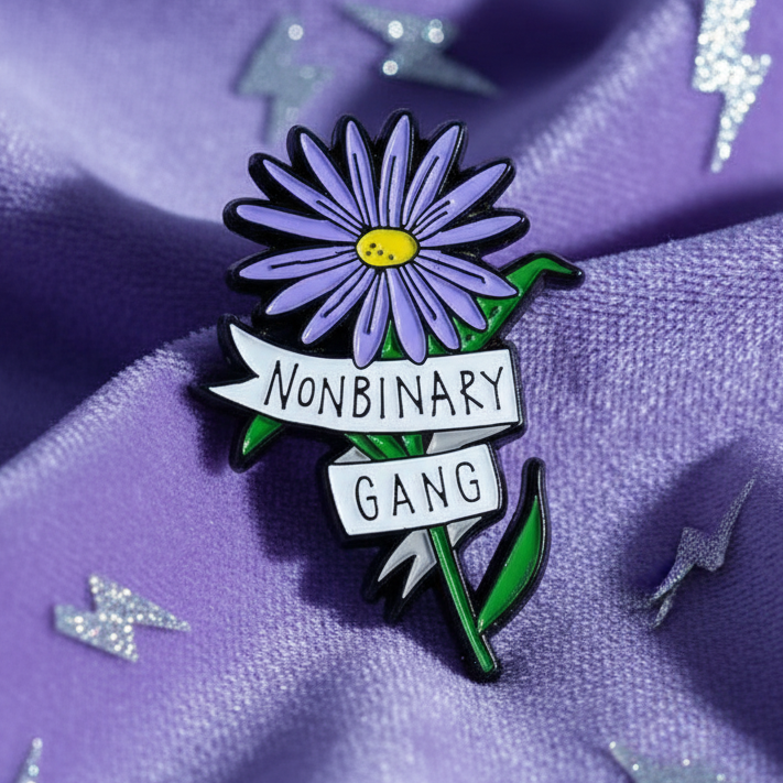Non-Binary Gang Enamel Pin: A vibrant purple daisy with a “NONBINARY GANG” banner, symbolizing pride and empowerment, resting on purple fabric with glittery accents. Perfect for expressing queer visibility and self-expression.