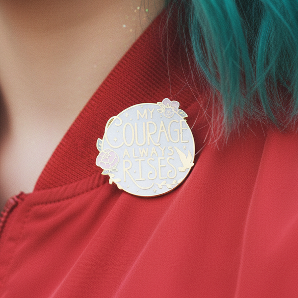 Close-up of a pastel enamel pin reading “MY COURAGE ALWAYS RISES” on a red garment