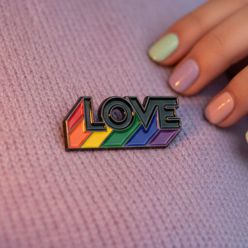 Love Is Love Rainbow Pride Enamel Pin on lavender knit fabric, featuring bold black LOVE letters over rainbow stripes. A hand with pastel-painted nails adds a touch of queer, empowering self-expression.