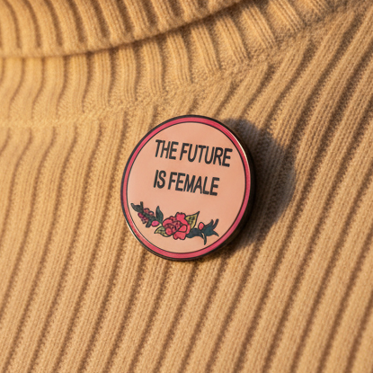 Round enamel pin reading “THE FUTURE IS FEMALE” with floral artwork on ribbed tan fabric