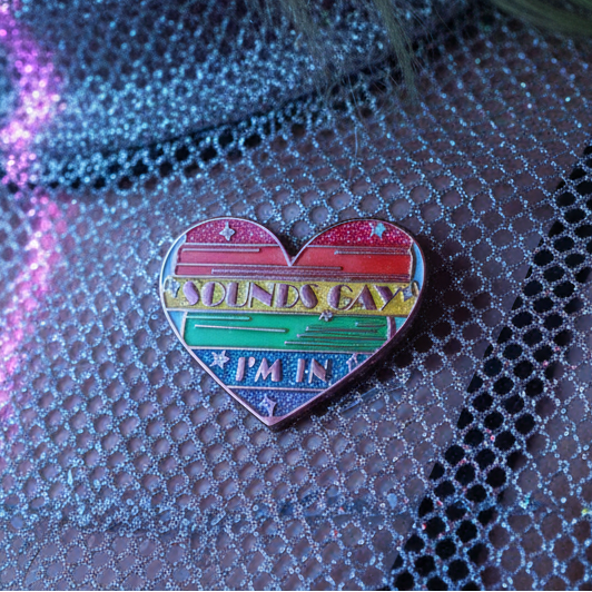 “Sounds Gay, I'm In” rainbow heart enamel pin on black fishnet fabric