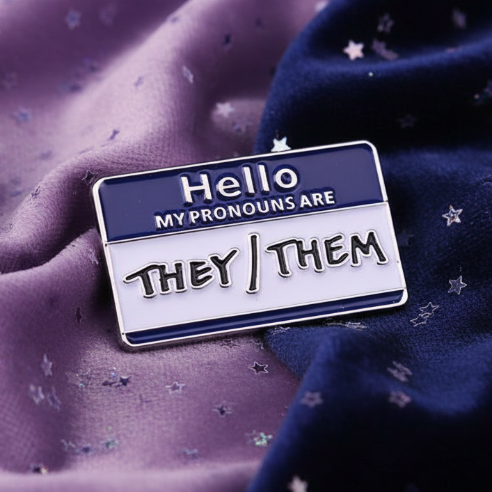 They/Them Pronouns Enamel Pin styled as a “Hello, my name is” badge. Dark blue top displays “Hello,” with “my pronouns are” beneath. White bottom declares “they/them.” Pin rests on purple fabric.