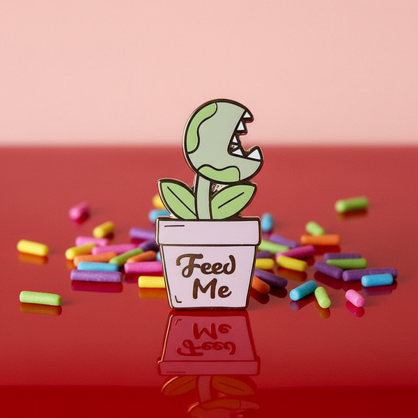 “Feed Me” enamel pin featuring a toothy potted carnivorous plant, surrounded by colorful candy