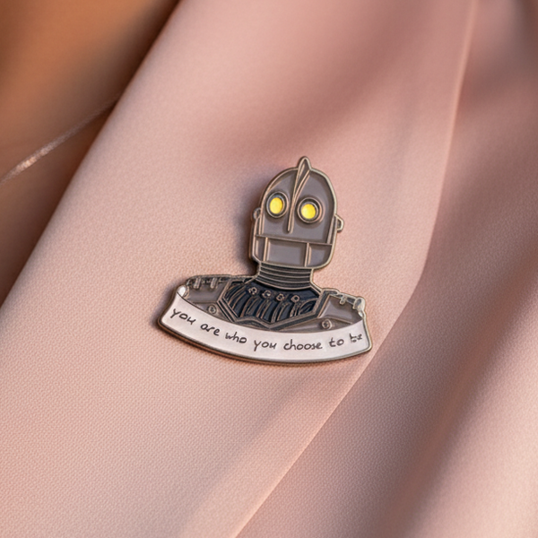 Close-up enamel pin of a robot with yellow eyes and “You are who you choose to be” text