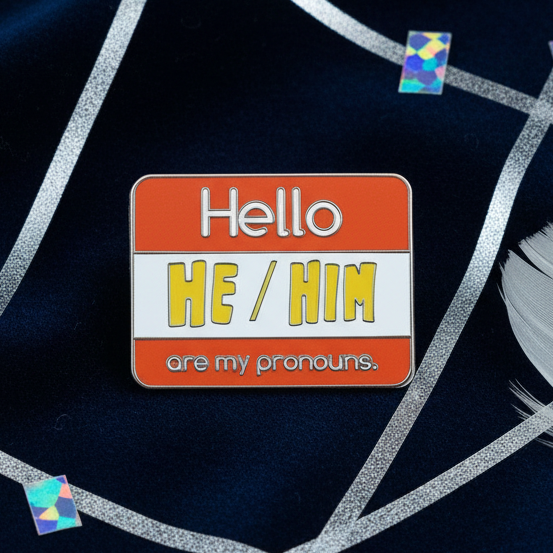 Enamel pin reading “Hello HE / HIM are my pronouns” on a black background