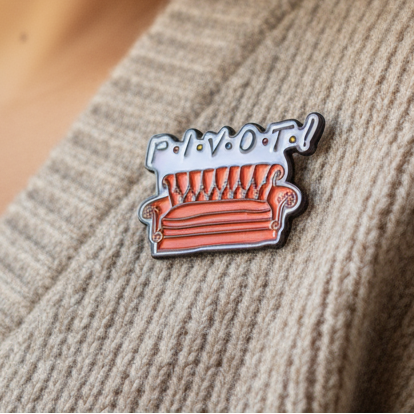 PIVOT enamel pin shaped like a red couch with white lettering on a sweater