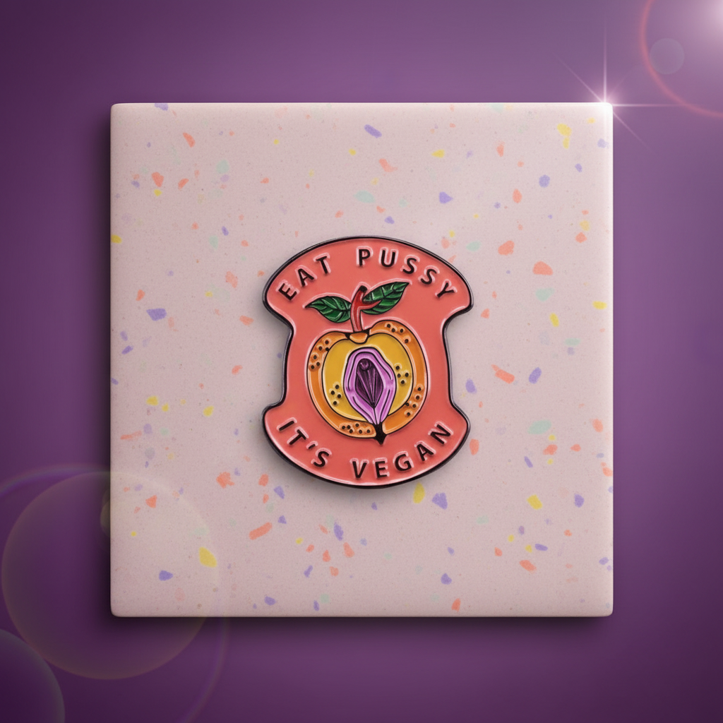 Close-up enamel pin reading “EAT PUSSY IT’S VEGAN,” with a vulva and fruit design