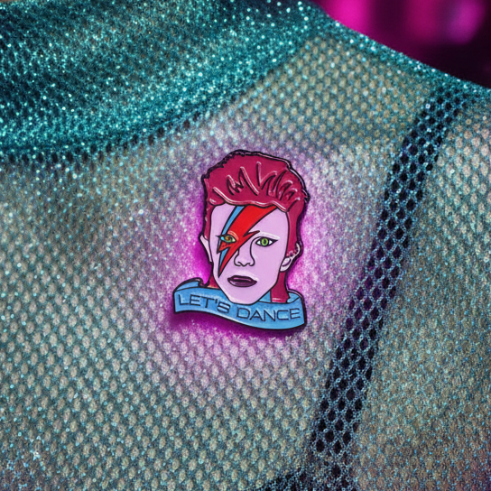Close-up of a Let's Dance enamel pin showing a pink-haired lightning-bolt-faced character