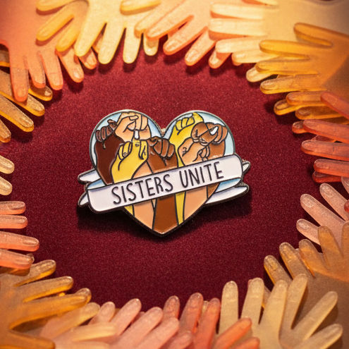 Sisters Unite Enamel Pin showcasing four diverse raised fists under a SISTERS UNITE banner, framed by orange and yellow glove shapes, embodying queer pride, solidarity, and empowerment.