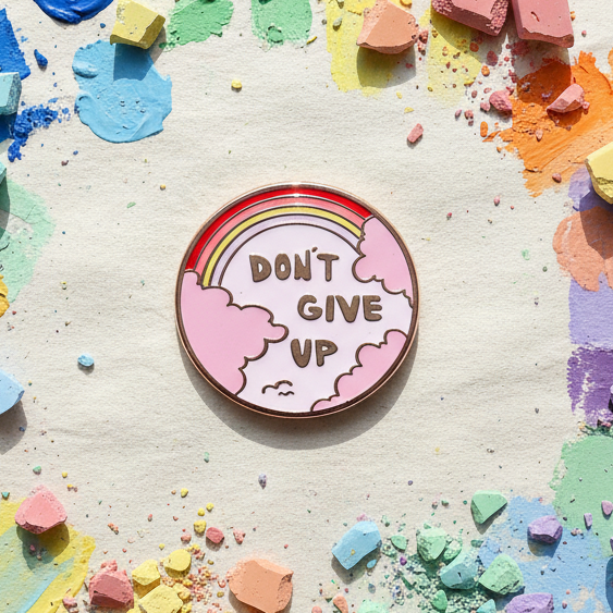 Round enamel pin reading “DON’T GIVE UP,” with a rainbow and pink clouds
