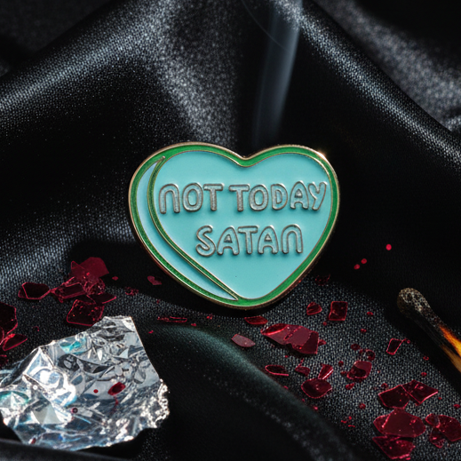 Turquoise-green heart-shaped Not Today Satan Enamel Pin with bold text, surrounded by a burnt matchstick and fragments, evokes fierce, prideful self-expression and empowerment in a queer lifestyle setting.