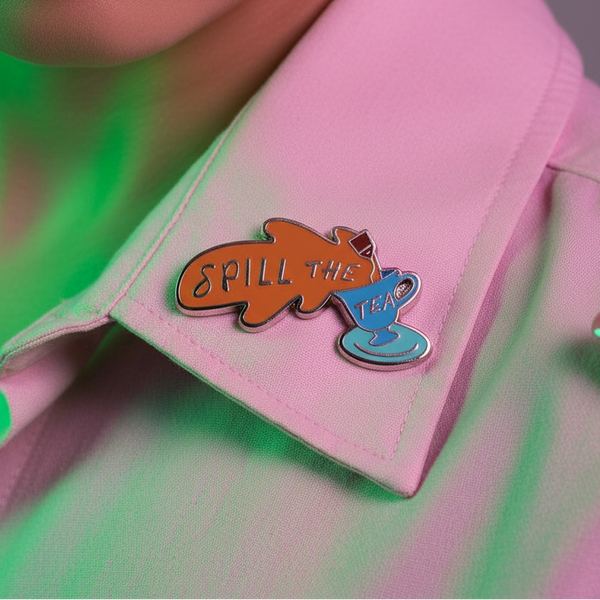 Close-up of a light pink collared shirt with the Spill The Tea enamel pin featuring a blue teacup and splash. Perfect for showcasing pride and queer empowerment with bold self-expression.