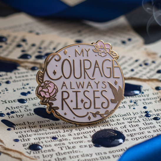 My Courage Always Rises Enamel Pin features gold-plated edges on lavender, adorned with a rose, bird, leaves, and stars, embodying pride and empowerment against yellowed book pages with blue ink splatters.