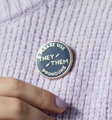 Round enamel pin reading “PLEASE USE THEY / THEM PRONOUNS” on lavender fabric