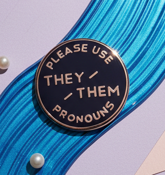 Close-up of a round enamel pin reading “PLEASE USE THEY–THEM PRONOUNS”
