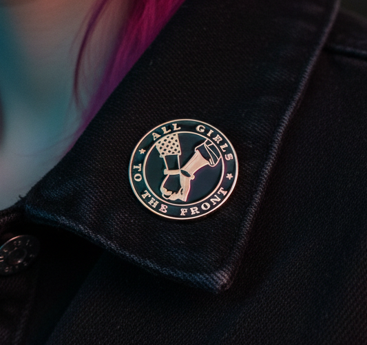 Close-up of an enamel pin reading “ALL GIRLS TO THE FRONT” on black fabric