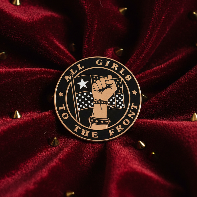 “All Girls to the Front” enamel pin with raised-fist artwork on burgundy studded fabric