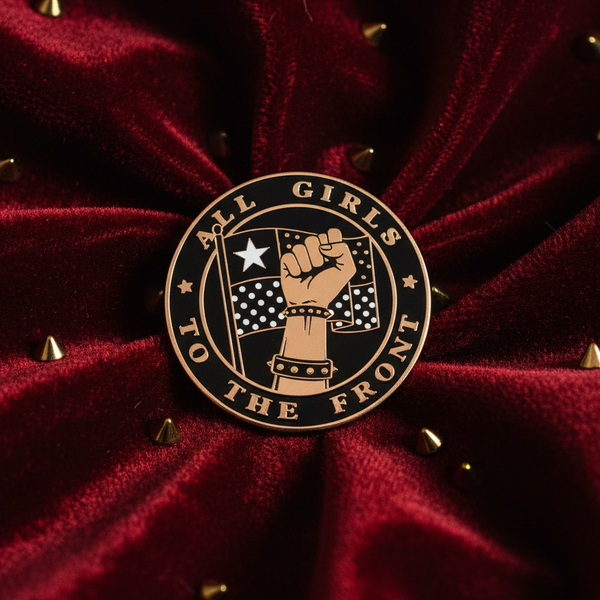 “All Girls to the Front” enamel pin with raised-fist artwork on burgundy studded fabric