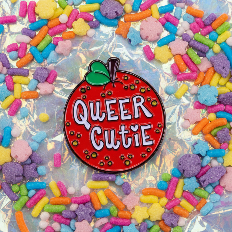Red apple-shaped enamel pin reading “QUEER CUTIE,” surrounded by colorful candy