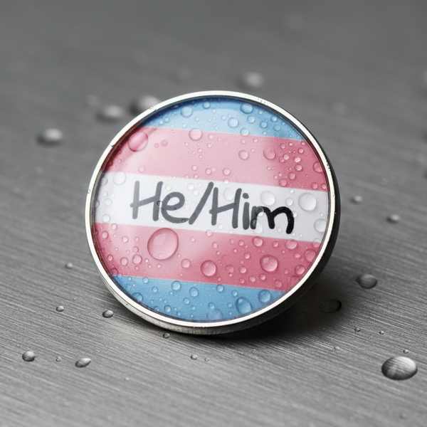 Close-up of a round He/Him enamel pin with trans pride flag stripes and water droplets