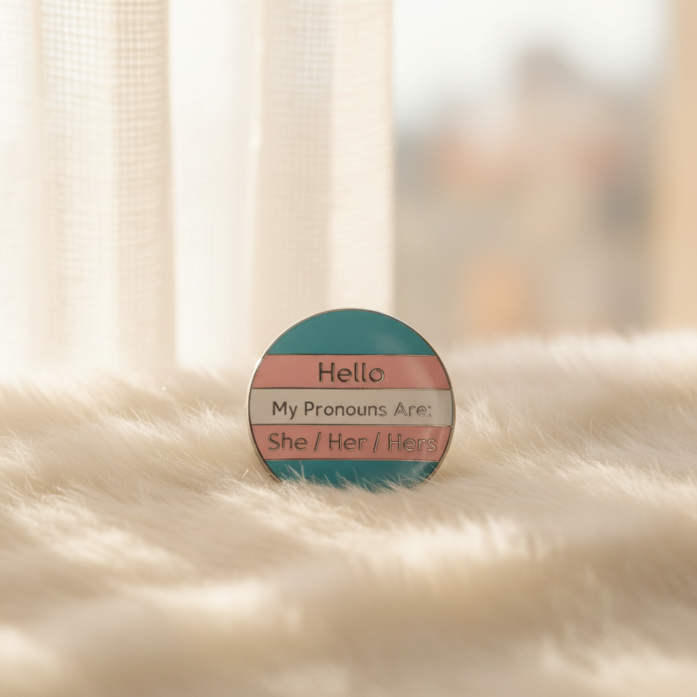 Round trans pride enamel pin reading “Hello My Pronouns Are She / Her / Hers”