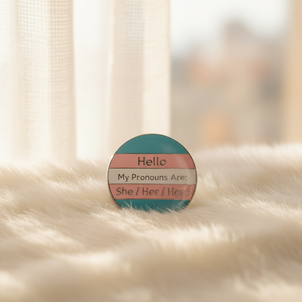 Round trans pride enamel pin reading “Hello My Pronouns Are She / Her / Hers”