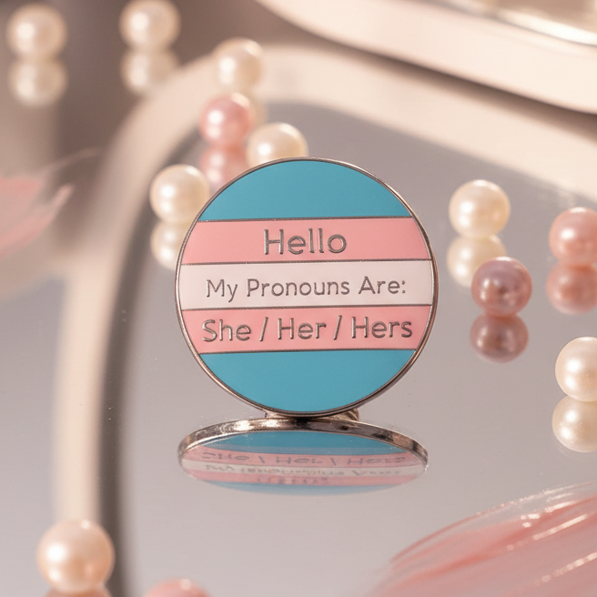 Round trans pride enamel pin reading “Hello My Pronouns Are: She / Her / Hers”