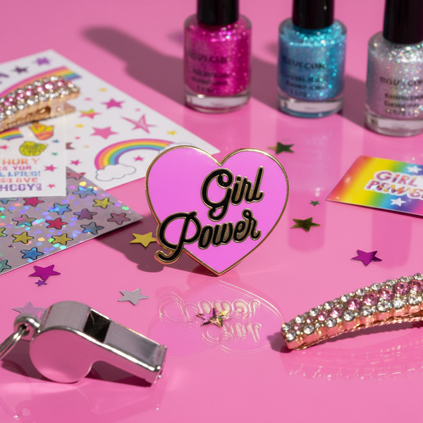 Pink heart enamel pin reading “Girl Power” on a glitter-covered pink display