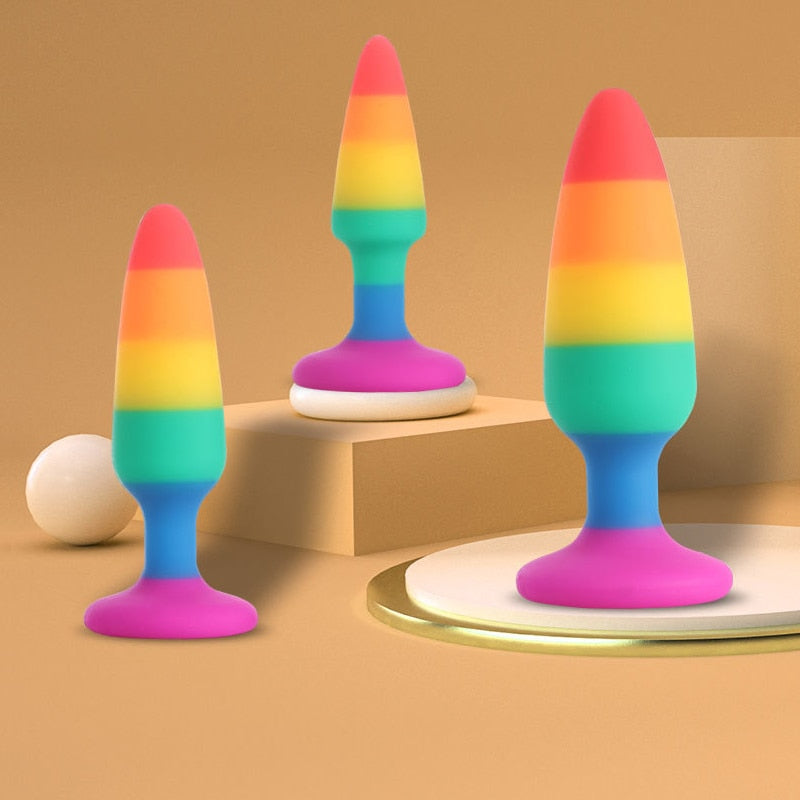 Three rainbow-striped butt plugs with blue stems and pink bases on tan display surfaces