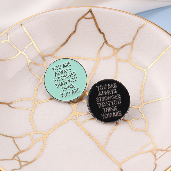 Two round enamel pins on a white, gold-crackled plate, mint and black, reading “YOU ARE ALWAYS STRONGER THAN YOU THINK YOU...