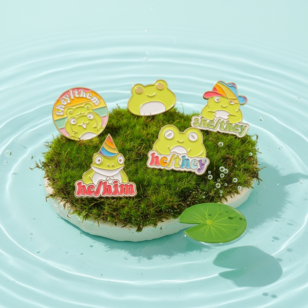 Four frog enamel pins reading “any pronouns,” “he/him,” “he/they,” and “she/they” on moss in water