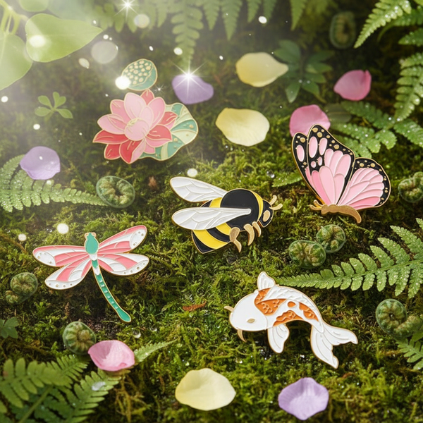 Five colorful enamel pins—a lotus, bee, butterfly, dragonfly, and koi—on moss and ferns