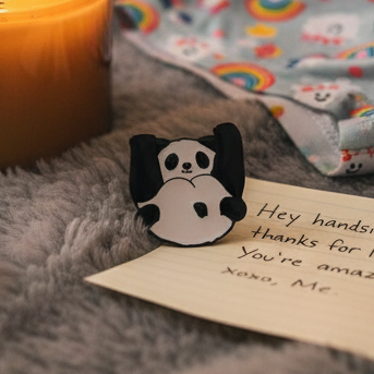Panda Bottom Enamel Pin displayed on a plush surface beside a loving note and a lit candle, accented by a vibrant rainbow scarf, embodying pride and self-expression.