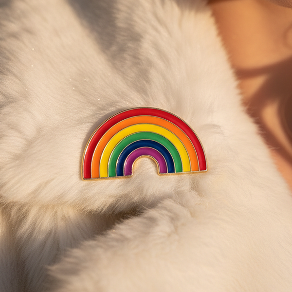 Close-up of a soft white faux-fur coat adorned with an LGBT Rainbow Enamel Pin on the lapel, symbolizing pride. The wearer exudes bold style with red lipstick, sparkly earring, and wavy blonde hair.