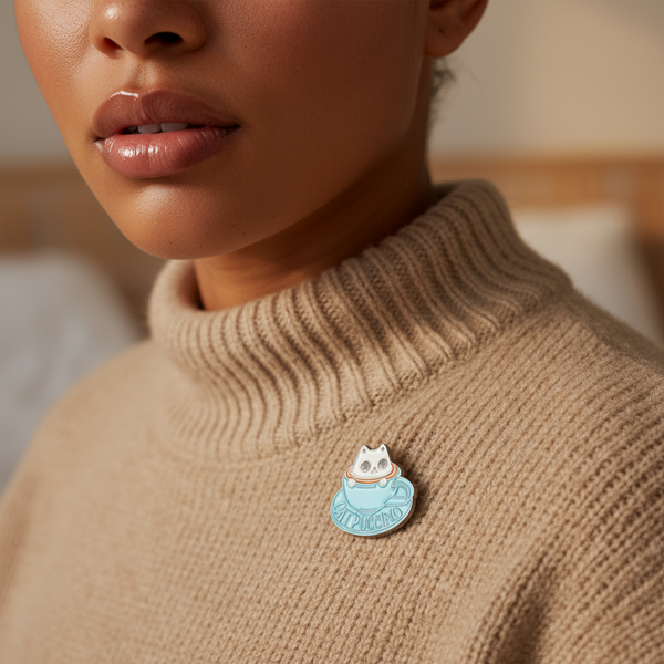 Close-up of a cat-shaped Catpuccino enamel pin on a tan ribbed sweater