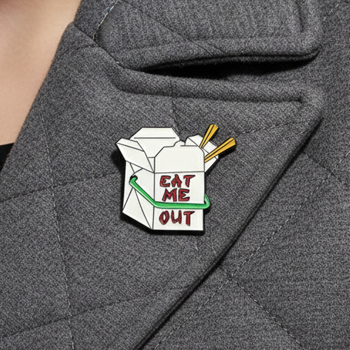 Close-up of an “EAT ME OUT” takeout carton enamel pin on a gray jacket