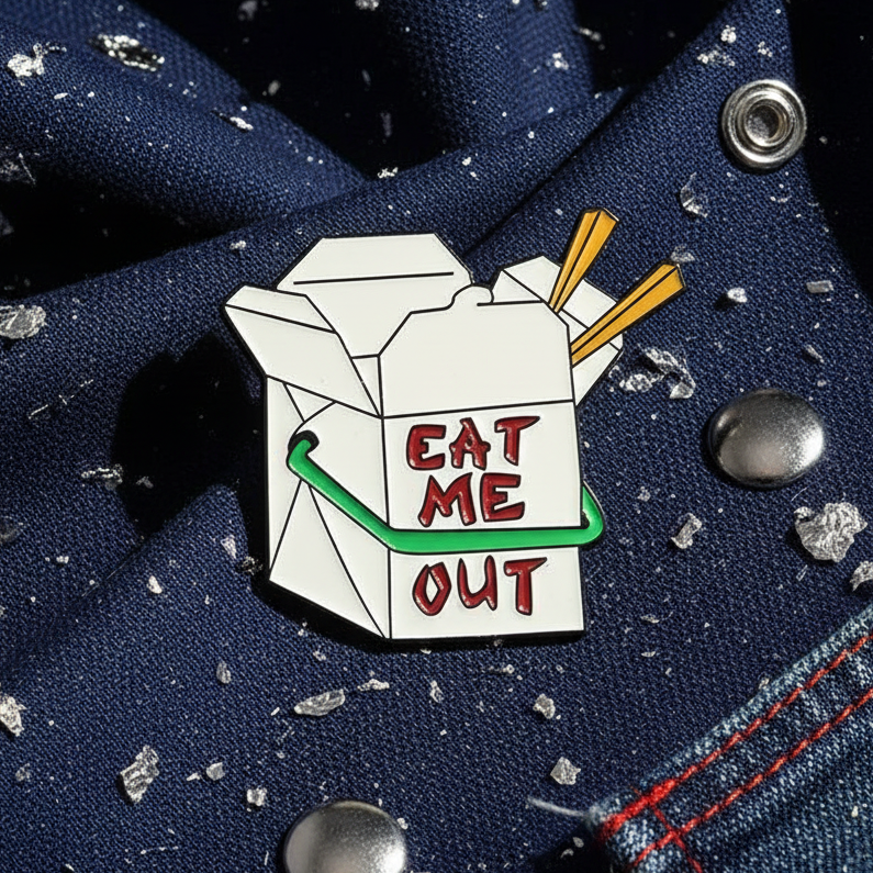 Close-up of an enamel pin shaped like a takeout box reading “EAT ME OUT”