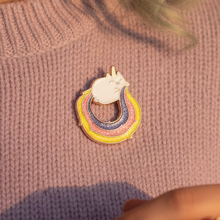 Close-up of Rainbow Kitty Enamel Pin featuring a stylized unicorn head with rainbow bands, worn on a lavender sweater. Pastel hair and rings subtly visible, embodying queer pride and self-expression.