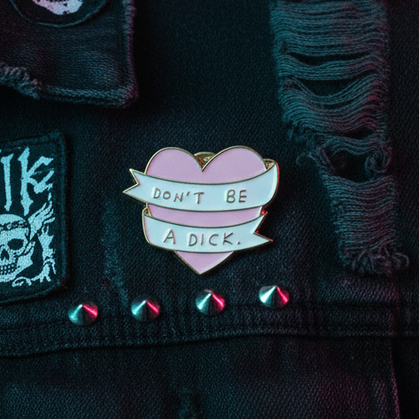 Heart-shaped enamel pin reading “DON’T BE A DICK” on a black denim jacket
