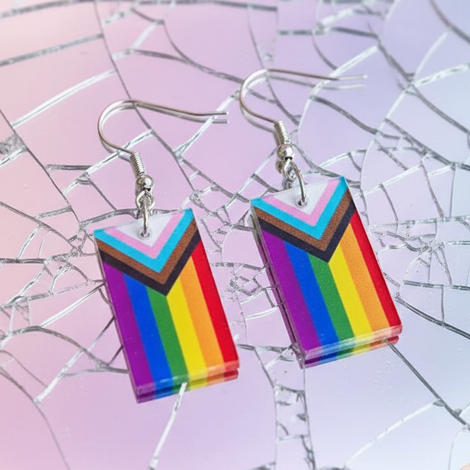 Pair of Progress Pride flag acrylic dangle earrings on a pink and lavender reflective surface