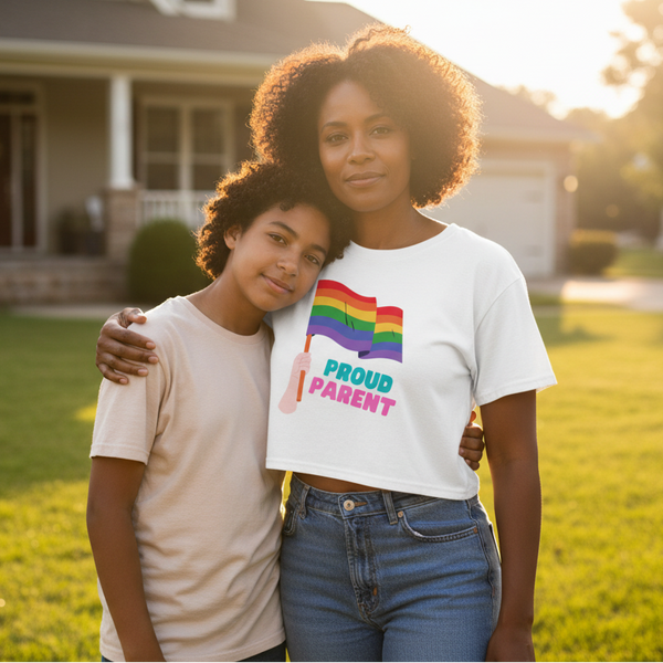 Adult and child pose together outdoors; the adult wears a white crop top reading “PROUD PARENT” with rainbow flags.