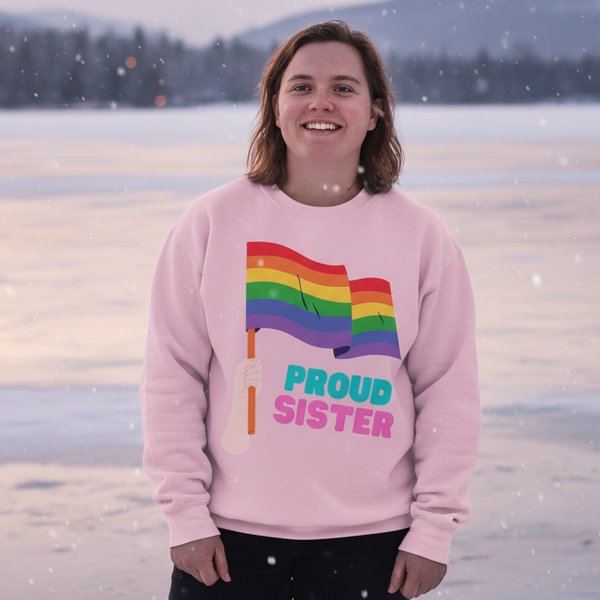 Smiling person wearing a pink sweatshirt reading “PROUD SISTER” with rainbow flags in falling snow