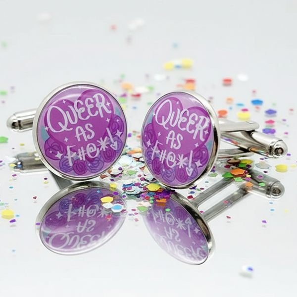 Pair of round purple “QUEER AS F#@*!” cufflinks with rose designs on a reflective surface