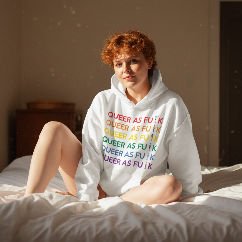 Person sitting on a bed in a white hoodie reading “QUEER AS FU*K” in rainbow lettering