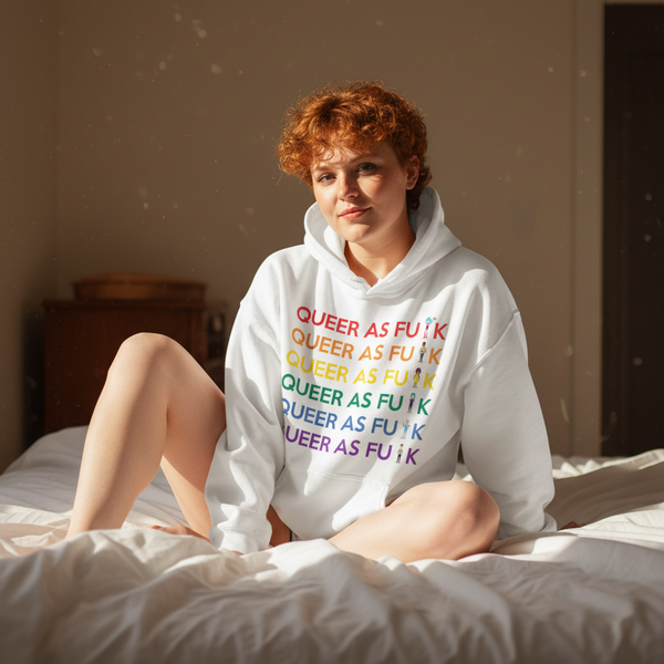 Person sitting on a bed in a white hoodie reading “QUEER AS FU*K” in rainbow lettering