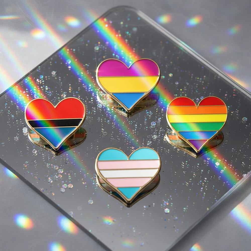 Four heart-shaped enamel pins with pride-style rainbow stripes on a glittery mirrored surface