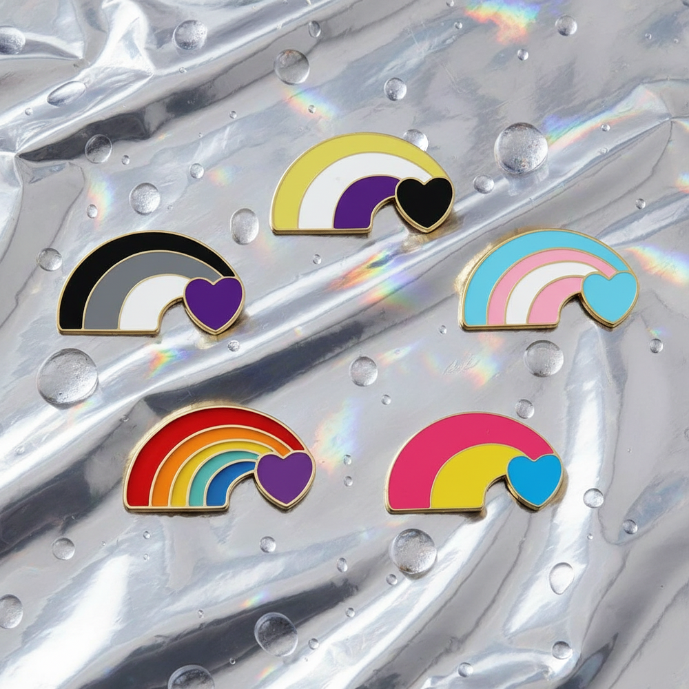 Flat lay of five rainbow enamel pins with heart accents on iridescent fabric