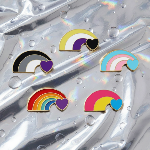 Flat lay of five rainbow enamel pins with heart accents on iridescent fabric