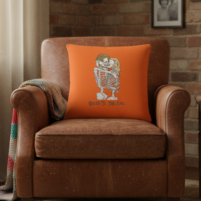 Orange "Queer til the end" pillow with two embracing skeletons on a brown leather armchair, crocheted blanket, brick wall